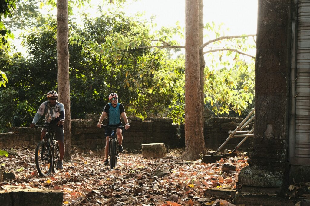 Cyclists riding beneath trees beside weathered Khmer temple ruins