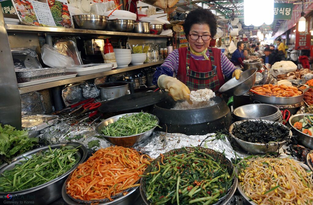 Smiling food vendor preparing rice beside colourful Korean dishes at Gwangjang Market in Seoul